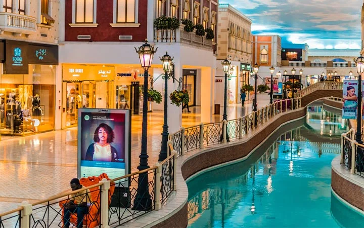 Villaggio Mall Qatar features an indoor canal, Venice-style bridges, and elegant street lamps. Shoppers stroll past digital ads and fashion stores beneath a ceiling painted like a blue sky with clouds, creating a unique shopping experience.
