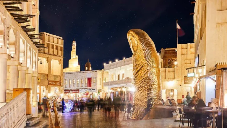 A lively, well-lit market square at night features a large, golden thumb sculpture surrounded by people—perfect for a family holiday in Qatar—with traditional Middle Eastern buildings and a minaret visible under the starry sky.