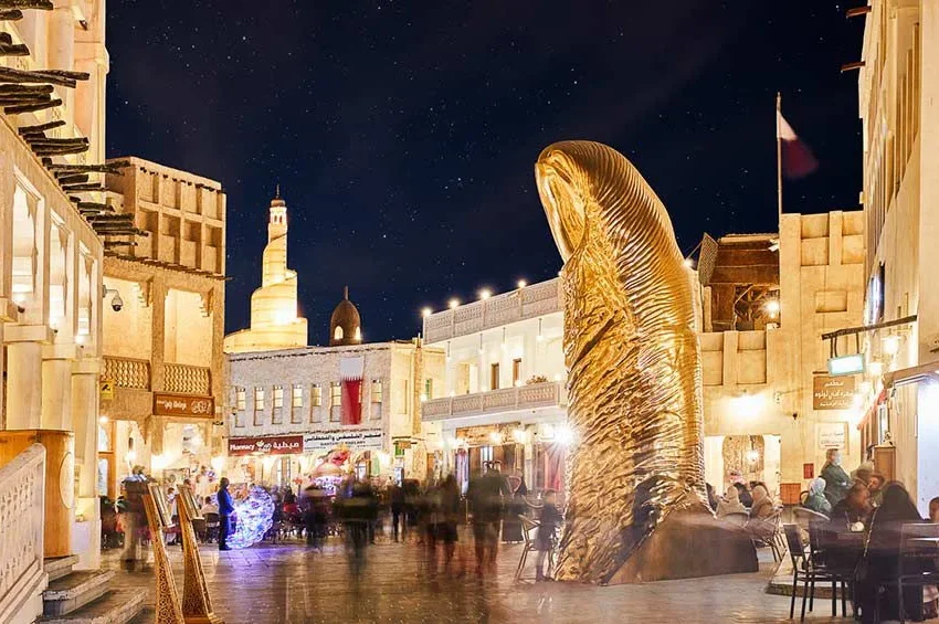 A lively, well-lit market square at night features a large, golden thumb sculpture surrounded by people—perfect for a family holiday in Qatar—with traditional Middle Eastern buildings and a minaret visible under the starry sky.