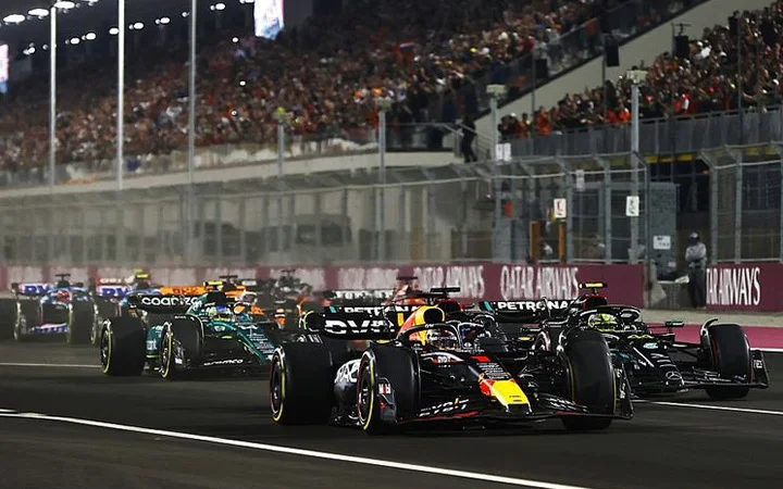 Several Formula 1 cars race closely together on a brightly lit track at night during the thrilling Formula 1 Qatar event, with grandstands full of spectators in the background and fencing lining the circuit.