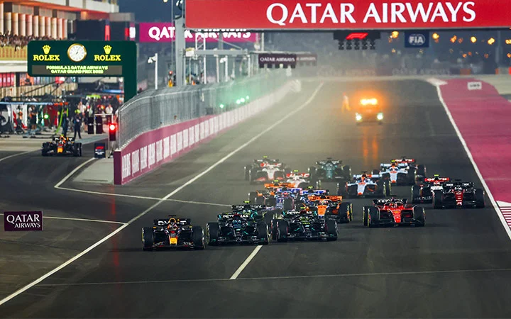 Formula 1 Qatar cars race off the starting line at night under bright lights at the Grand Prix, with the Qatar Airways banner overhead and pit crews visible in the background.