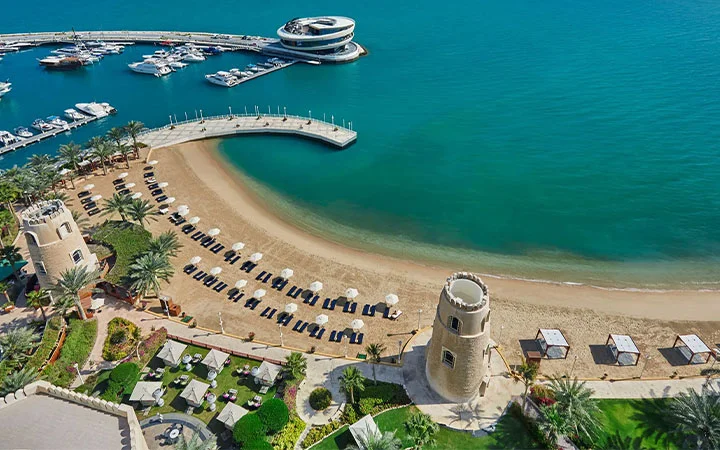 Aerial view of Doha Beach with sun loungers, umbrellas, palm trees, and two stone towers beside turquoise water. Luxury yachts are docked near the Four Seasons Doha, with a modern circular building on the water in the background.