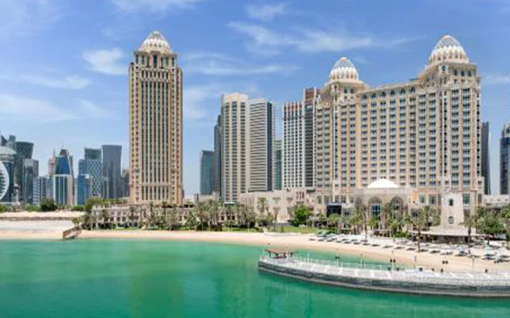 A view of modern skyscrapers and luxury hotels like Four Seasons Doha along the sandy Doha Beach with turquoise water under a clear blue sky in a stunning cityscape.