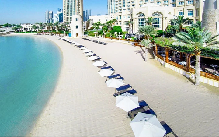 Aerial view of a sandy Doha Beach lined with white umbrellas and lounge chairs beside turquoise water, bordered by palm trees and modern buildings, showcasing the luxury hotel ambiance of Four Seasons Doha under a clear blue sky.