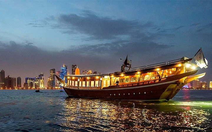 A traditional wooden dhow boat adorned with lights floats on calm water at dusk, offering a serene Dhow Cruise dinner experience as the modern city skyline glows in the background beneath a partly cloudy sky.