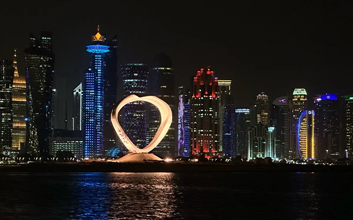 Night view of Doha’s skyline featuring illuminated skyscrapers and the glowing golden Lusail Plaza Tower, with shimmering reflections on the water—a perfect scene for a Dhow Cruise dinner under the full moon.