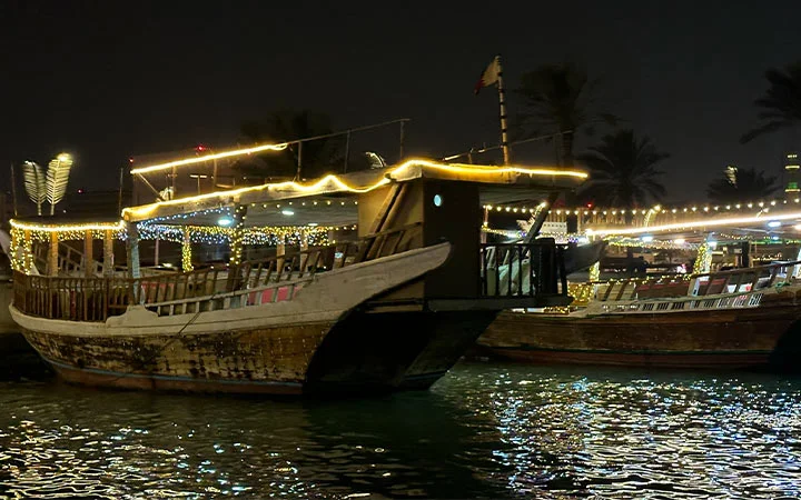 Traditional wooden boats decorated with string lights float on the water at night, offering a magical Dhow Cruise experience under the Full Moon, with illuminated reflections shimmering and palm trees in the background.