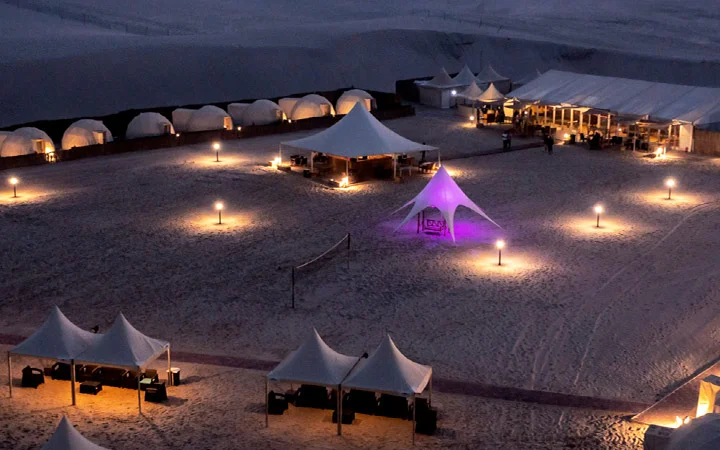 A nighttime view of a desert campsite under the glow of the Full Moon Overnight Camping experience, featuring white tents, canopies, and warm lights on the sand, with a central tent lit in purple and a large tented area in the background.