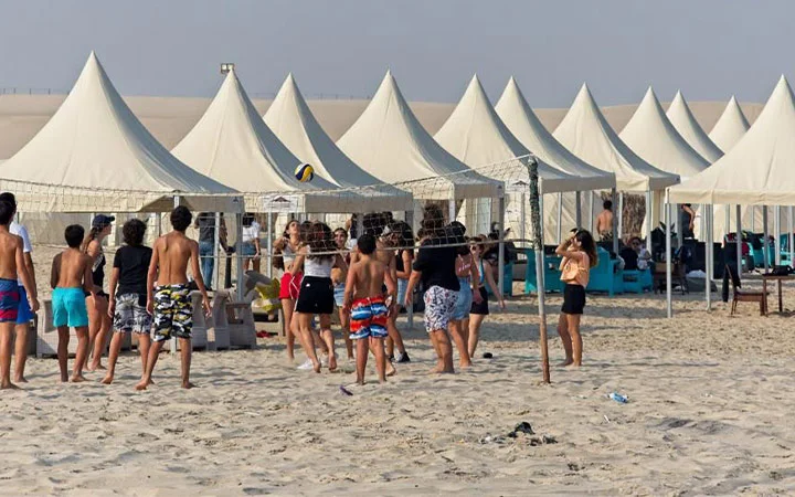 A group of people play beach volleyball on sandy ground in front of several white tents, perfect for Full Moon Overnight Camping. Some people watch from inside the tents, enjoying the sunny and clear weather.