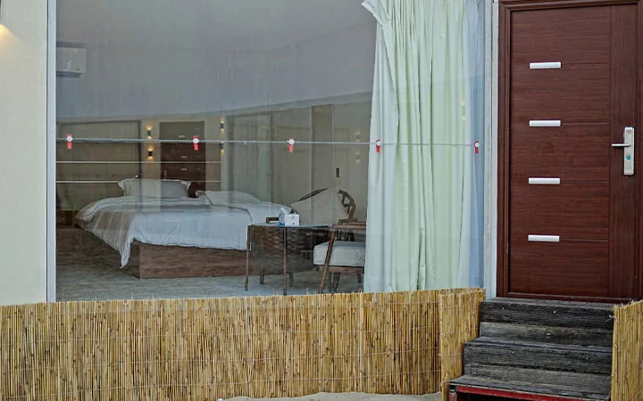 A hotel room with a large glass window reveals a neatly made bed, a small table, and chair—perfect for relaxing after Full Moon Overnight Camping. Outside, bamboo panels and steps lead to a wooden door with silver handles and accents.