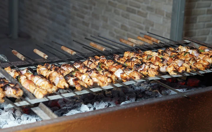 Several skewers of marinated meat and vegetables are grilling over hot coals during a Full Moon Overnight Camping trip, with a stone wall in the background. The skewers are evenly spaced and partially charred, indicating they’re being cooked.
