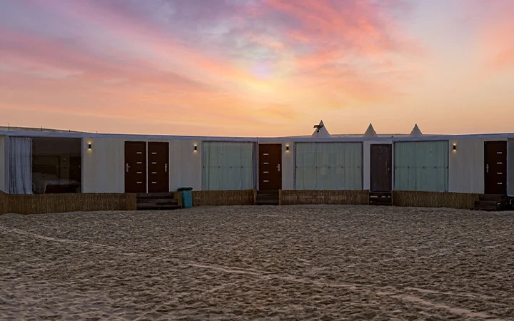 A row of modern tent cabins with wooden doors and lights stands on sandy ground under a colorful sunset sky, perfect for Full Moon Overnight Camping. The area in front is open and spacious.