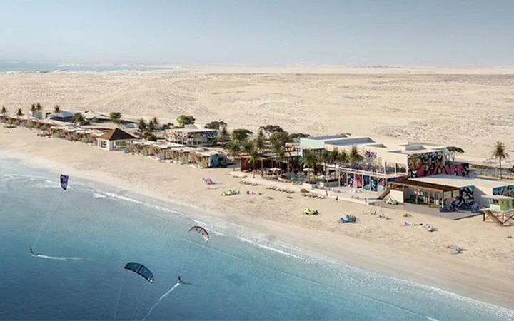 A sandy beach with colorful modern buildings, palm trees, and people kitesurfing in the blue ocean at Fuwairit Kite Beach. The scene shows a blend of desert and beach, with lounge chairs and beachgoers relaxing near the water.