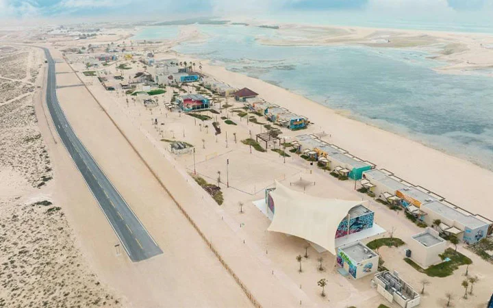 Aerial view of Fuwairit Kite Beach, Qatar, with a straight road on the left, rows of colorful buildings, palm trees, and shaded structures along the sandy beach—perfect for kite surfing beside the calm, shallow sea.