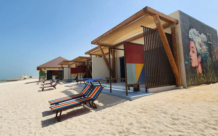 Colorful beach huts with modern geometric designs and wall art face the sandy beach at Fuwairit Kite Beach in Qatar, with striped lounge chairs arranged outside and perfect conditions for kite surfing under a clear blue sky.