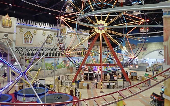 An indoor amusement park featuring a Ferris wheel, roller coaster tracks, colorful lights, and themed decorations, with various attractions and people visible in the background.