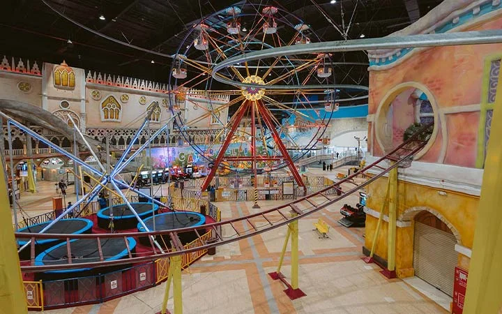 An indoor amusement park with a Ferris wheel, roller coaster tracks, and various bright, colorful structures and arcade machines in the background. The area is spacious and decorated with whimsical, carnival-themed designs.