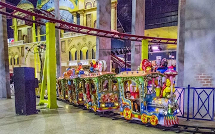 A colorful indoor amusement park train ride with cartoon-themed carriages travels along tracks, with children and lights visible, set against a backdrop of faux building facades and a roller coaster overhead.
