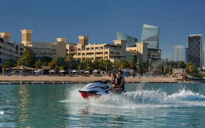 Two people ride a jet ski on calm blue water near a sandy beach at the Grand Hyatt Beach resort, with modern and traditional-style buildings and palm trees in the background under a clear sky.
