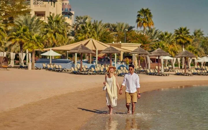 A couple walks hand in hand along the shoreline of Grand Hyatt Beach, with lounge chairs, sunshades, and palm trees visible in the background under a clear sky.