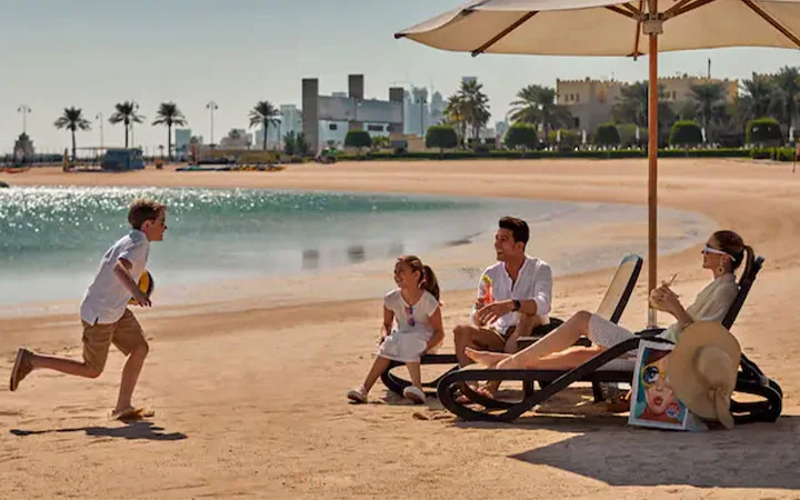 A family relaxes on a sandy beach near calm water at a luxury hotel; a boy runs toward a man, woman, and girl seated on lounge chairs under a large umbrella, with palm trees and buildings in the background.