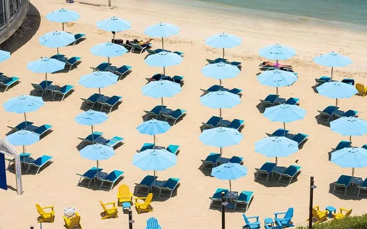 Rows of blue beach umbrellas and lounge chairs are neatly arranged on the sandy shores of West Bay Beach near the water, with a few yellow chairs in the foreground and the Hilton Doha hotel nearby.