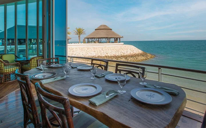 A wooden dining table set with plates, glasses, and napkins overlooks the beach at West Bay through large windows. Outside, palm trees and thatched-roof gazebos dot the sandy shore beneath Hilton Doha’s blue sky views.