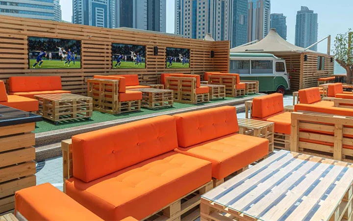 Outdoor lounge at Hilton Doha with orange cushioned pallet sofas and tables, large TVs on a wooden fence showing sports, and a retro van in the background—all set against the city skyline near West Bay Beach.