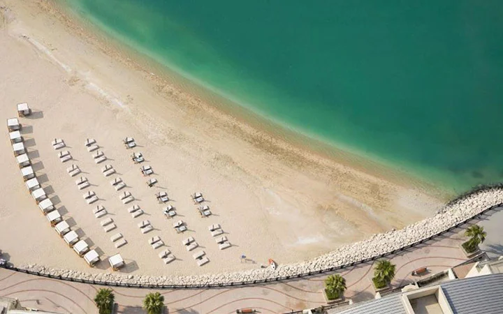 Aerial view of Pearl Beach at Hilton Doha, featuring a sandy shore lined with white lounge chairs and cabanas in a curve by clear turquoise water, bordered by a palm-lined walkway—an inviting scene at this luxurious Doha hotel.