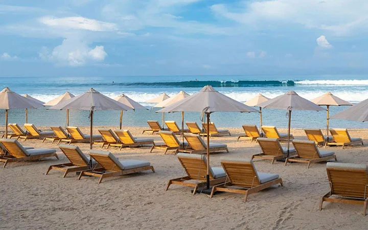 Wooden lounge chairs with white cushions and beige umbrellas are arranged on the sandy shores of Pearl Beach at Hilton Doha, facing the ocean’s gentle waves under a cloudy blue sky.