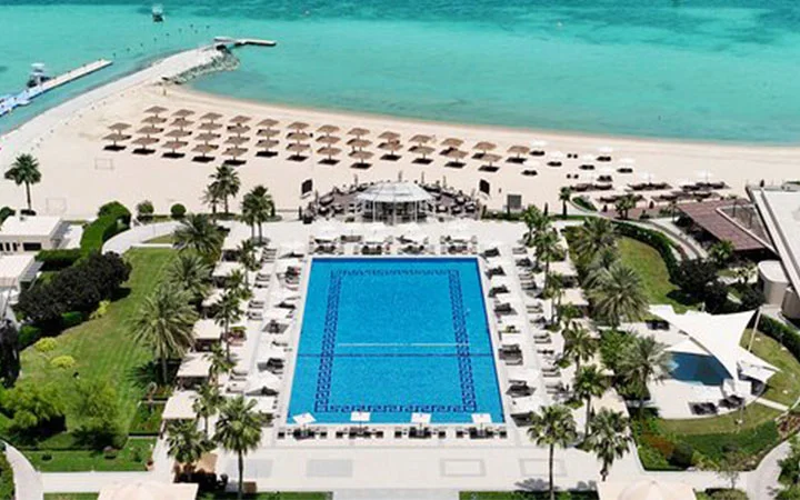 Aerial view of Hilton Doha resort with a large rectangular pool, lounge chairs, palm trees, and umbrellas on the sandy Pearl Beach along turquoise ocean water.