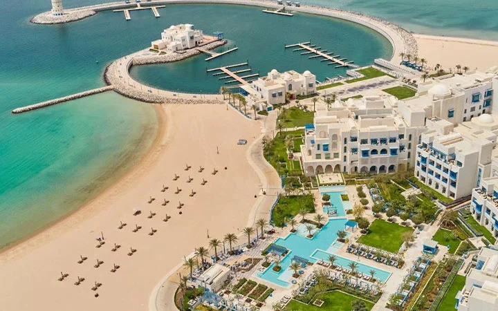 Aerial view of Hilton Salwa Beach, a luxurious beach resort with white buildings, multiple pools, palm trees, and a private sandy beach lined with loungers beside turquoise waters and a marina with docks extending into the sea.