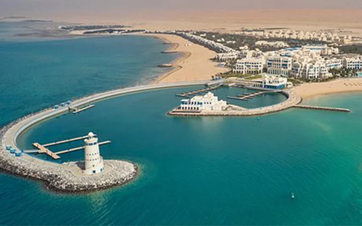 Aerial view of Hilton Salwa Beach Resort with a curved pier, two white lighthouses, and buildings surrounded by turquoise water and sandy beaches, set against a desert backdrop.