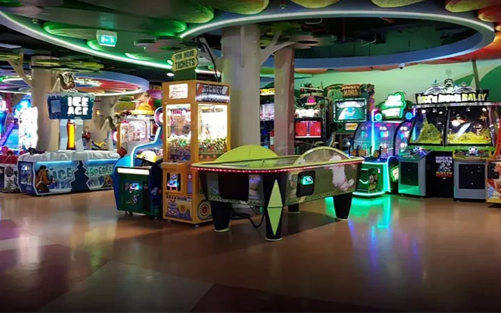 A brightly lit arcade with various colorful game machines, including an air hockey table in the center and ticket redemption games on the left, creating a lively and playful atmosphere.