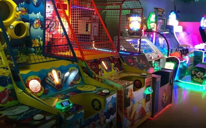A row of colorful arcade game machines, including basketball hoops and various light-up games, is illuminated by vibrant neon lights in a lively indoor gaming center.