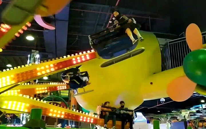 Children ride a colorful indoor amusement park ride shaped like an airplane, with bright yellow parts and lights illuminating the scene. The ride appears to be moving quickly, causing motion blur.