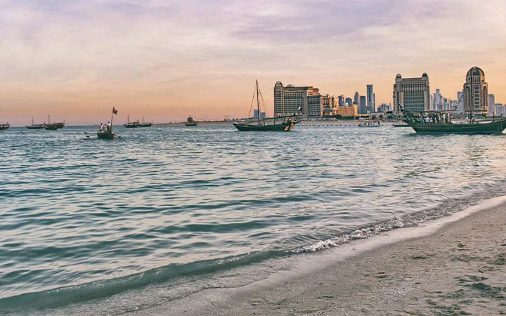 Calm sea with gentle waves on the sandy shore of Katara Beach, traditional boats floating on the water, and Doha’s modern city skyline with tall buildings in the background at sunset.