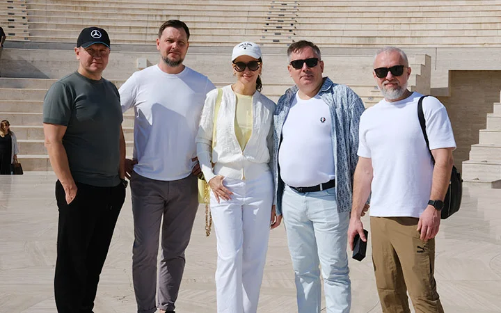 Five adults stand together outdoors on pale stone steps at Katara Village Tours, posing for a group photo. Four men and one woman, all in casual light-colored clothing and sunglasses, smile at the camera in bright sunlight.