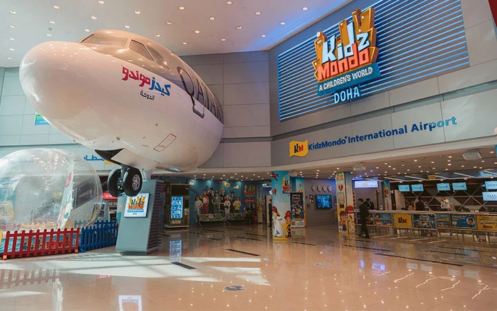 A large model airplane is mounted above the entrance to KidzMondo International Airport at KidzMondo Doha, a children’s play center with colorful signs and bright lighting.