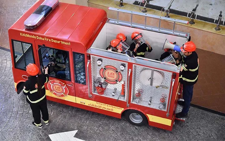 Five children dressed as firefighters with red helmets stand around and inside a small red fire truck marked KidzMondo Doha Fire Department, preparing for an activity in an indoor setting.