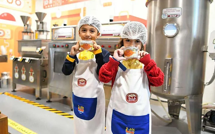 Two children wearing hairnets and aprons stand in a factory-like setting, smiling and holding packaged food products. Large industrial machines and bright decor are visible in the background.