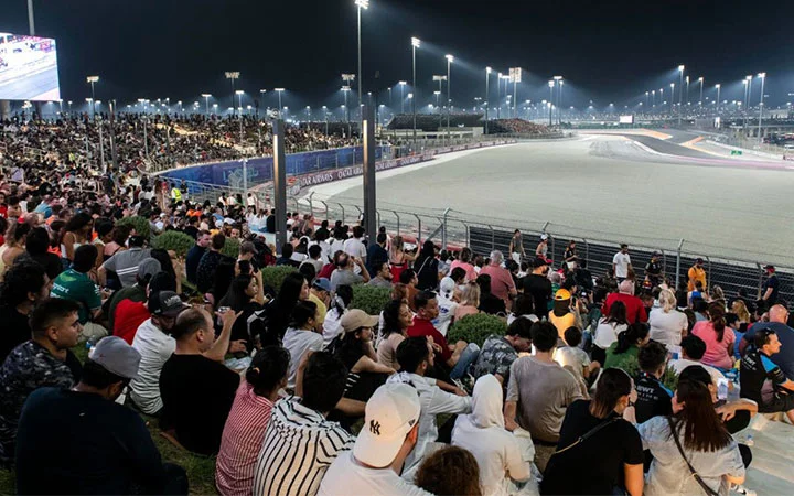 A large crowd of spectators sits on grassy seats beside the racetrack at night, with the exclusive Lusail Hill Lounge offering premium views under bright stadium lights. The stands are packed as the track curves through the background.