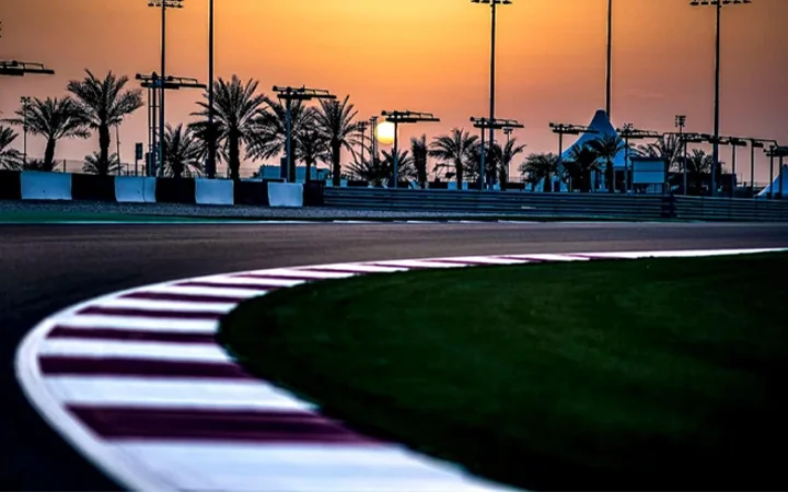A racing track curves into the distance at sunset near Lusail Hill Lounge, with palm trees and floodlights lining the background and the sun low on the horizon, creating a warm, dramatic sky.