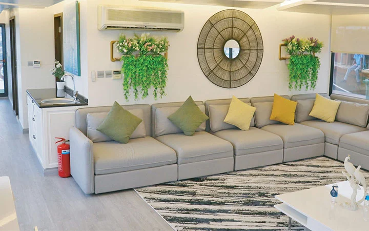Modern living room on a Private House Boat Qatar, featuring a long gray sectional sofa, colorful throw pillows, striped rug, wall art, plants, round mirror, air conditioner, and fire extinguisher near the kitchenette. Bright and neatly decorated space.