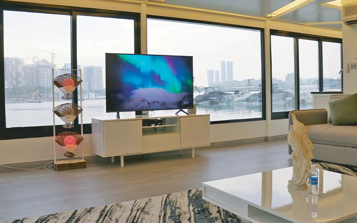 A modern living room with large windows overlooking a cityscape and water, inspired by a Private House Boat Qatar. A TV displaying northern lights sits on a white stand, beside a colorful floor lamp. A sofa and coffee table with decor complete the space.