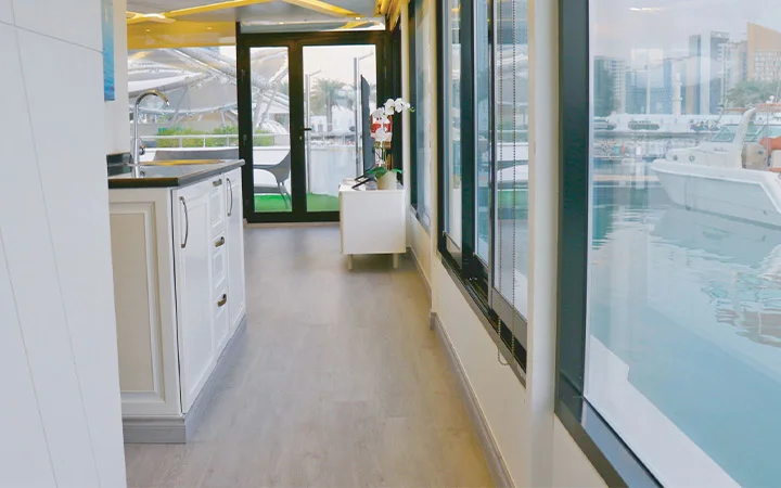 Modern Private House Boat Qatar interior with light wood flooring, white kitchen cabinetry, and large windows. Enjoy waterfront views of the marina, boats, and nearby buildings from the comfort of this stylish retreat.