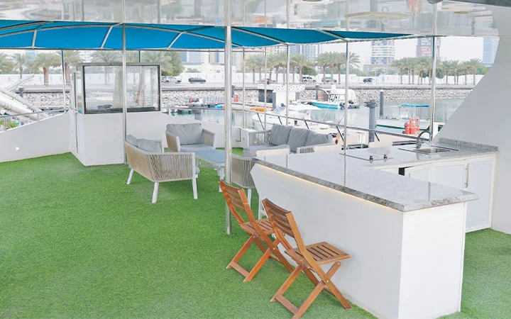 A yacht deck on a Private House Boat Qatar features artificial grass flooring, a marble-topped bar, wooden chairs, cushioned sofas, and a blue canopy, all overlooking a marina with boats and city buildings in the background.