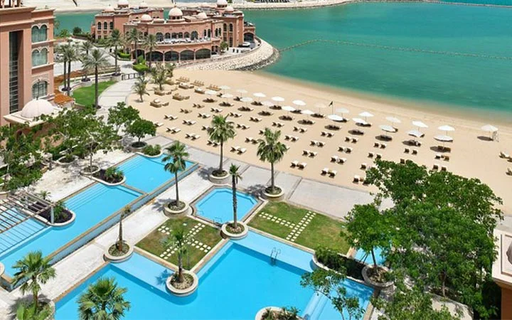 A resort beachfront at Marsa Malaz Kempinski with rows of lounge chairs and umbrellas on the sand, several turquoise pools surrounded by palm trees, and a large building overlooking a calm, blue sea.