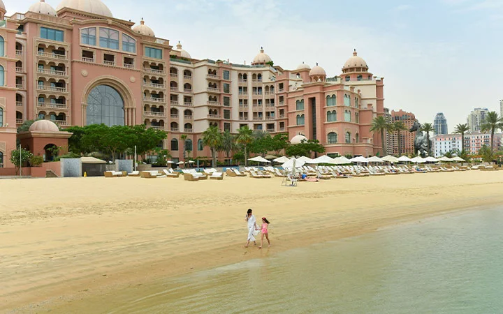 Two people stroll along a sandy beach near calm water, in front of the luxurious Marsa Malaz Kempinski hotel, its domes and balconies rising behind rows of beach chairs and umbrellas, with city buildings in the background.