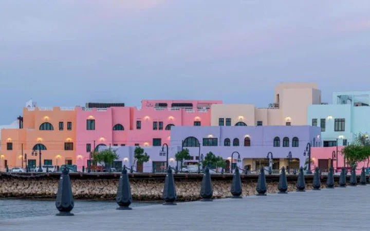 A row of pastel-colored buildings in pink, purple, blue, and beige stands by the waterfront, with lamp posts and trees along the paved promenade under a soft, early evening sky.